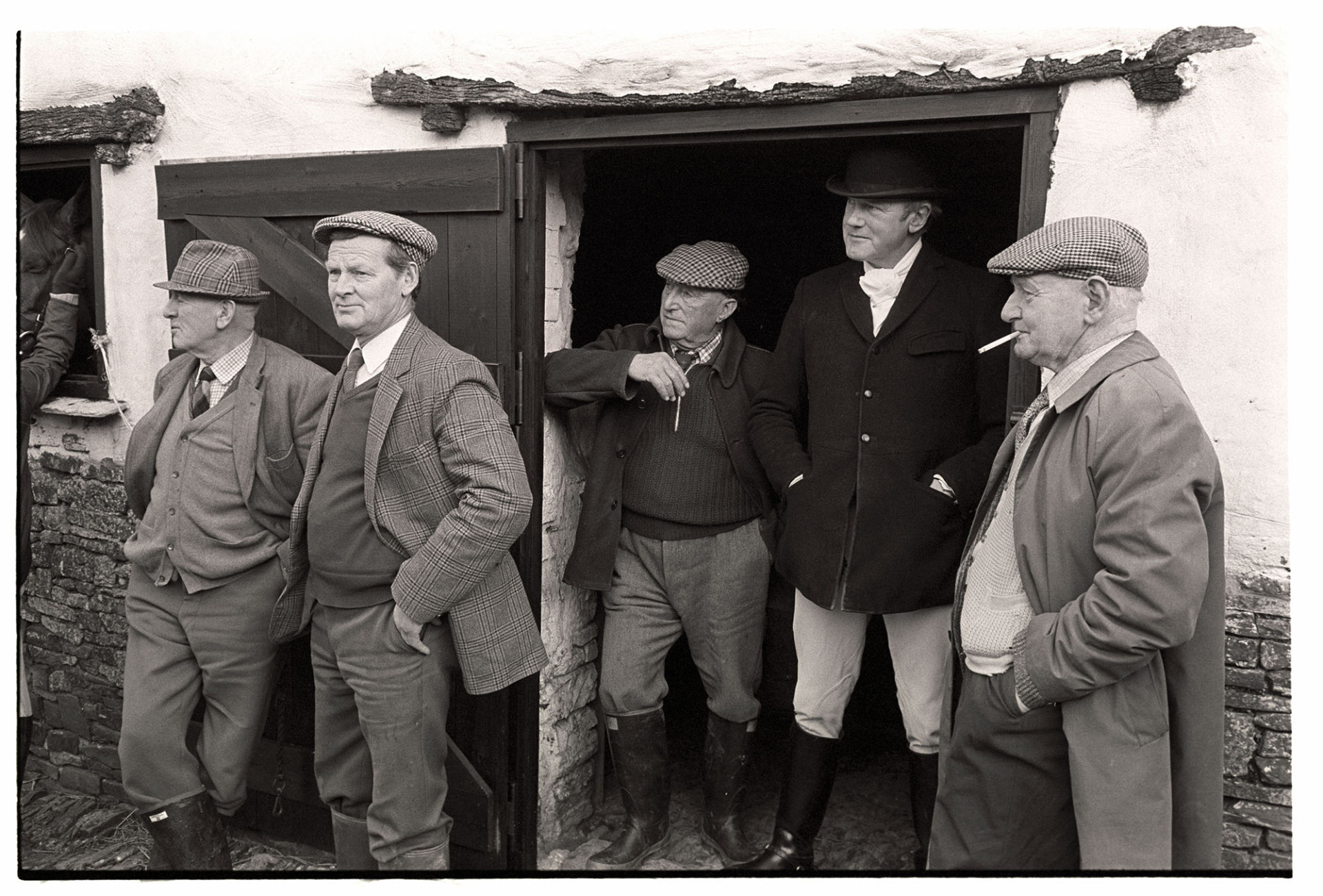 Hunt meet at house, followers and rider standing looking at meet, hunting clothes. 
[Five men stood around a doorway at a hunt meet at Hackwells, Dolton. One of the men is smoking a cigarette and another is dressed in riding or hunting clothes.]