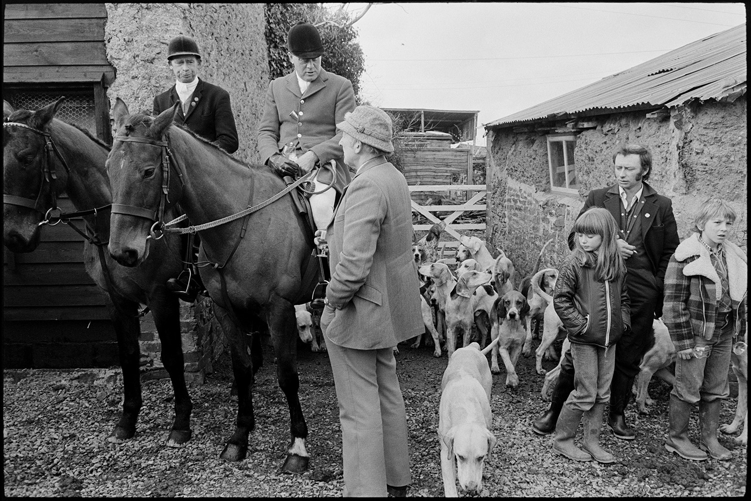 Hunt meet at house, followers and riders waiting, master, hounds setting off through village.
[Hunt Master mounted on a horse and another mounted rider in the farmyard of Hackwells, Dolton talking to a man. A pack of hounds and  a man and two children are with them by a barn.]