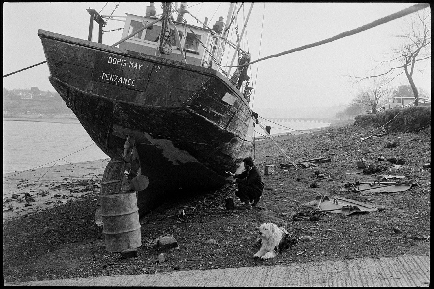 Beached fishing boat, repairs being carried out. 
[A man painting the fishing boat 'Doris May' which is moored on the bank of the River Torridge at Bideford. Bideford Long Bridge, also known as Bideford Old Bridge, is visible in the background. A dog is with the man.]
