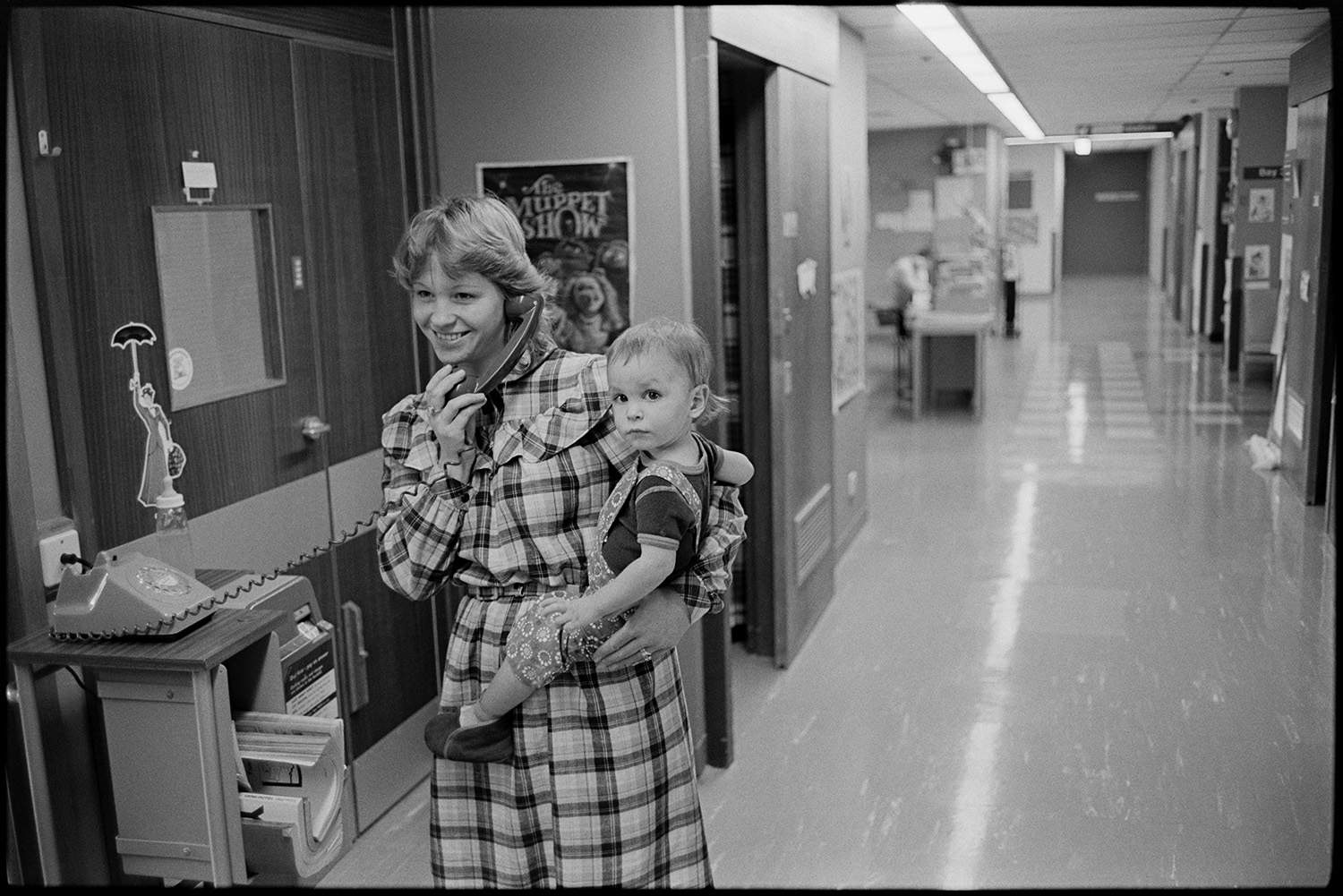 Hospital children's ward, women mothers with children in wards. Woman on telephone. 
[A woman holding a baby making a telephone call from the children's ward at Barnstaple General Hospital. The ward can be seen in the background and a poster advertising The Muppet Show is pinned to the wall behind her.]