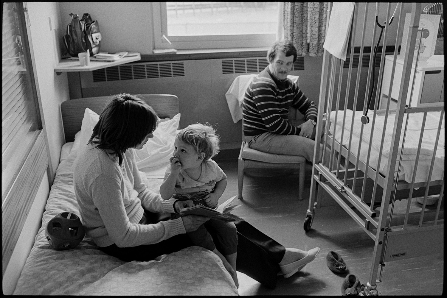 Hospital children's ward, women mothers with children in wards. Woman on telephone. 
[A woman, child and man on the children's ward at Barnstaple General Hospital. The woman and child are sat on a bed reading a book. A cot is next to them.]