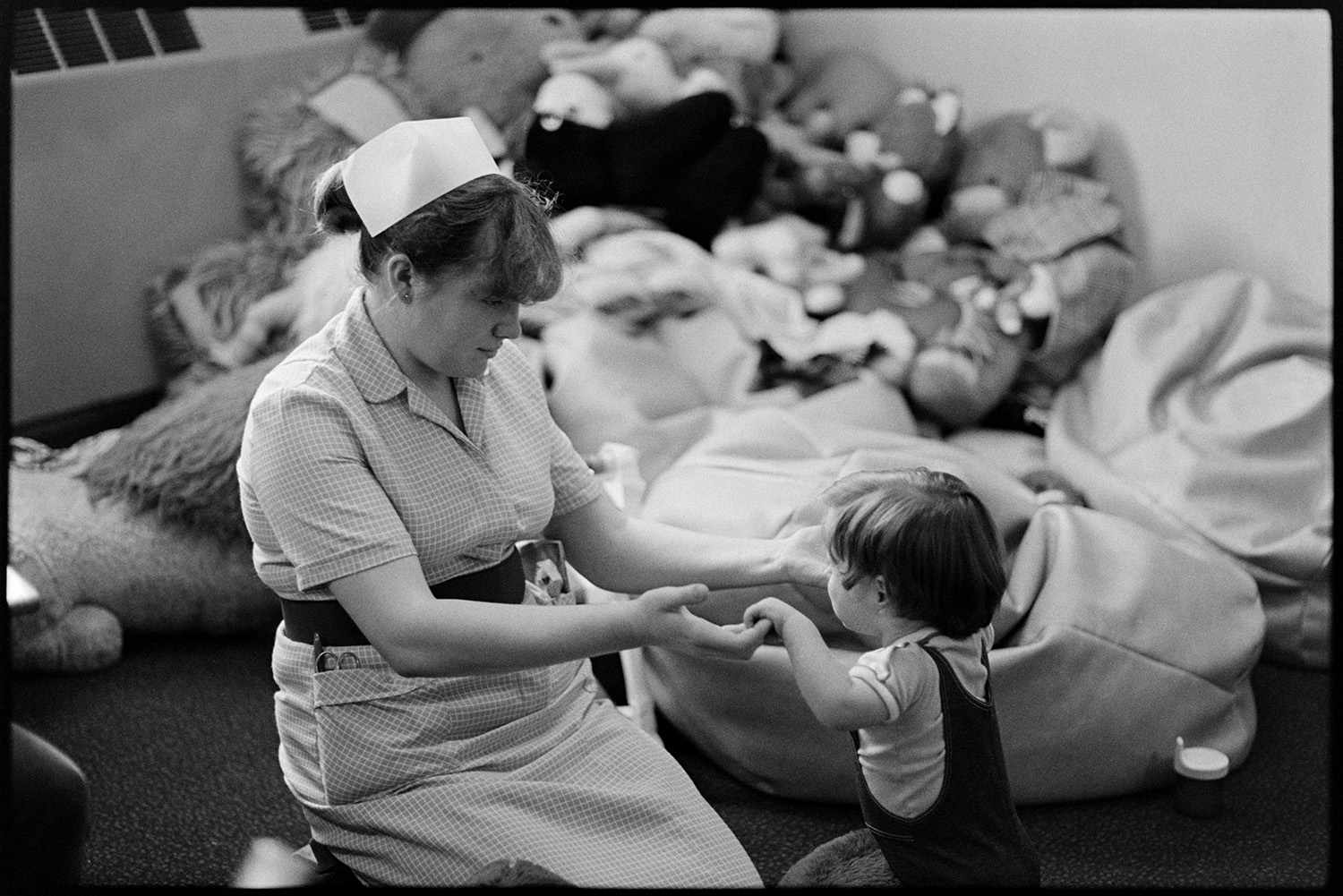 Hospital children's ward, nurses, mothers with children in wards, Sister at ward, cuddly toys. 
[A nurse playing with a young child in the Children's Ward at Barnstaple general Hospital. A pile of bean bags and soft toys can be seen in the background.]