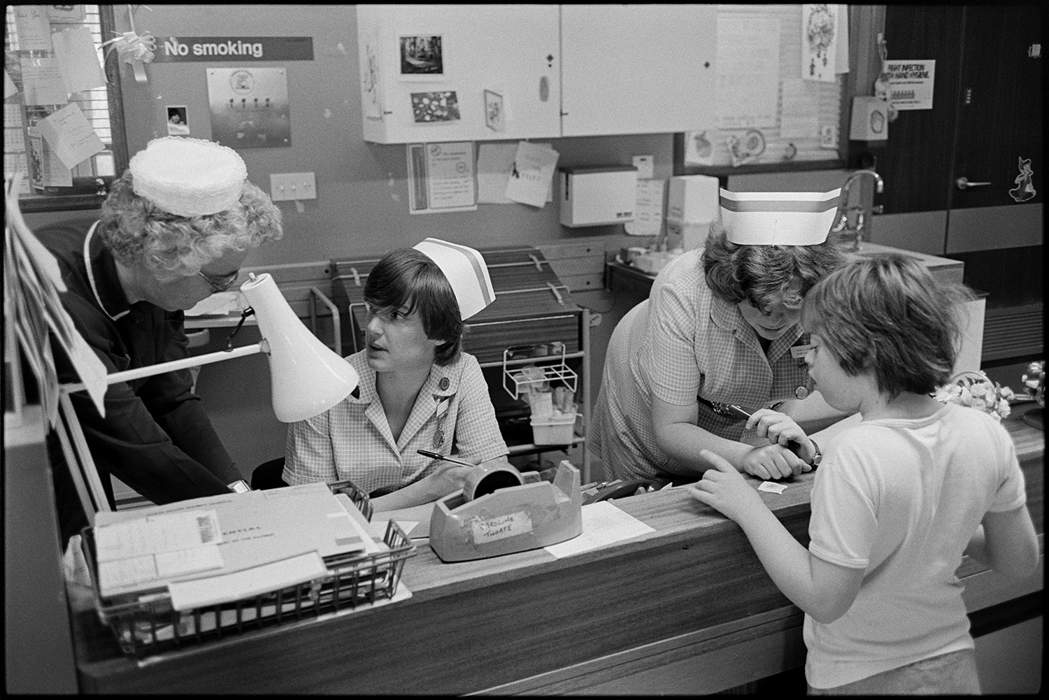 Hospital children's ward, nurses, mothers with children in wards, Sister at ward, cuddly toys. 
[The ward Sister and nurses talking at the reception of the Children's Ward at Barnstaple General Hospital. A child is at the reception desk.]