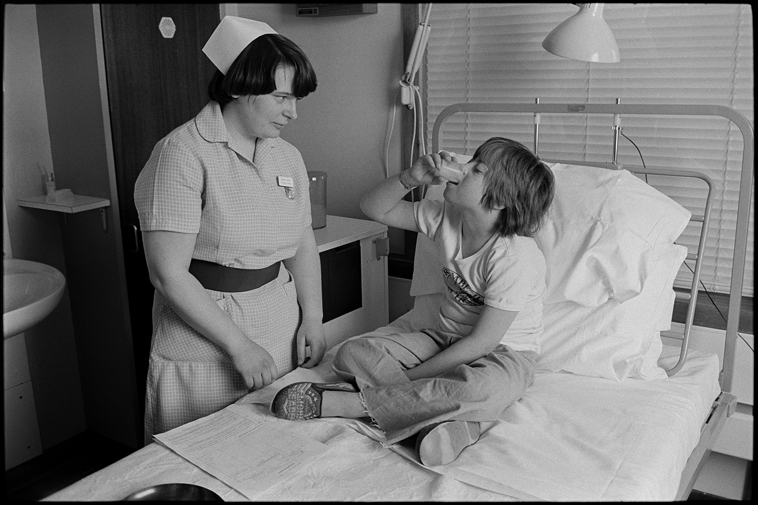 Hospital children's ward, eating, father visiting daughter in bed, Sister and Doctor, bed making. 
[A nurse giving a child medication in a hospital bed on the Children's Ward at Barnstaple General Hospital.]