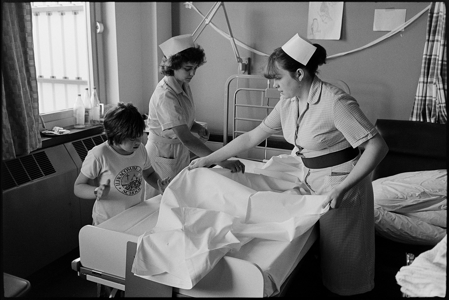 Hospital children's ward, eating, father visiting daughter in bed, Sister and Doctor, bed making. 
[Two nurses and a child changing bedding  in the Children's Ward at Barnstaple General Hospital.]