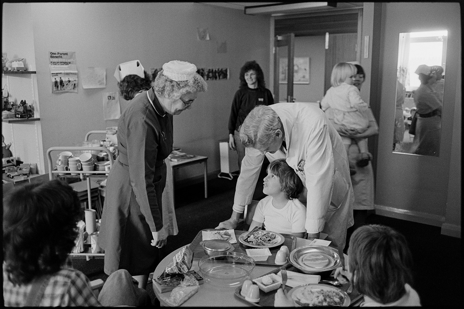 Hospital children's ward, eating, father visiting daughter in bed, Sister and Doctor, bed making. 
[The ward Sister and a doctor talking to a child having lunch at the Children's Ward at Barnstaple General Hospital. Other children and women are in the background.]
