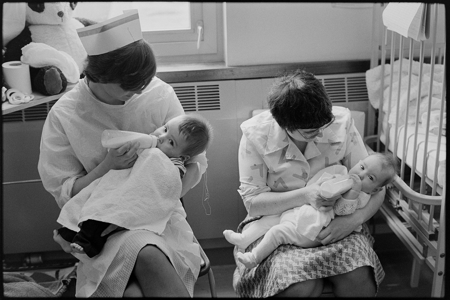Hospital children's ward, nurses, woman, mother feeding babies, doctor examining child, teacher. 
[Two women, one of which is a nurse, feeding bottles of milk to babies on the Children's Ward at Barnstaple General Hospital.]