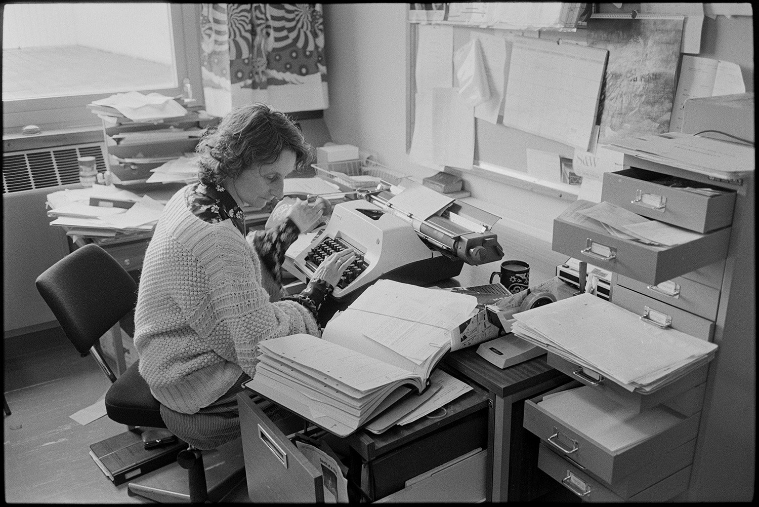Hospital. Doctor's secretary at her desk typing. Woman. <br />
[A doctor's secretary typing up notes on a typewriter at Barnstaple General Hospital. Various papers on are on her desk and a filing cabinet with open drawers is next to the desk.]