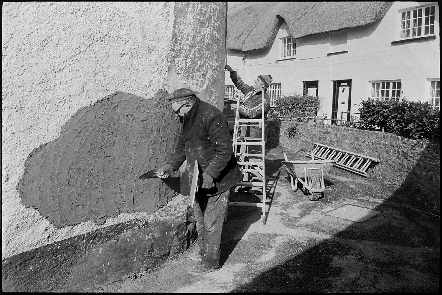 Derek Marden and Clifford Palmer repairing the cob wall of a house in North Street, Dolton. Ladders and wheelbarrow are in the street next to them.
