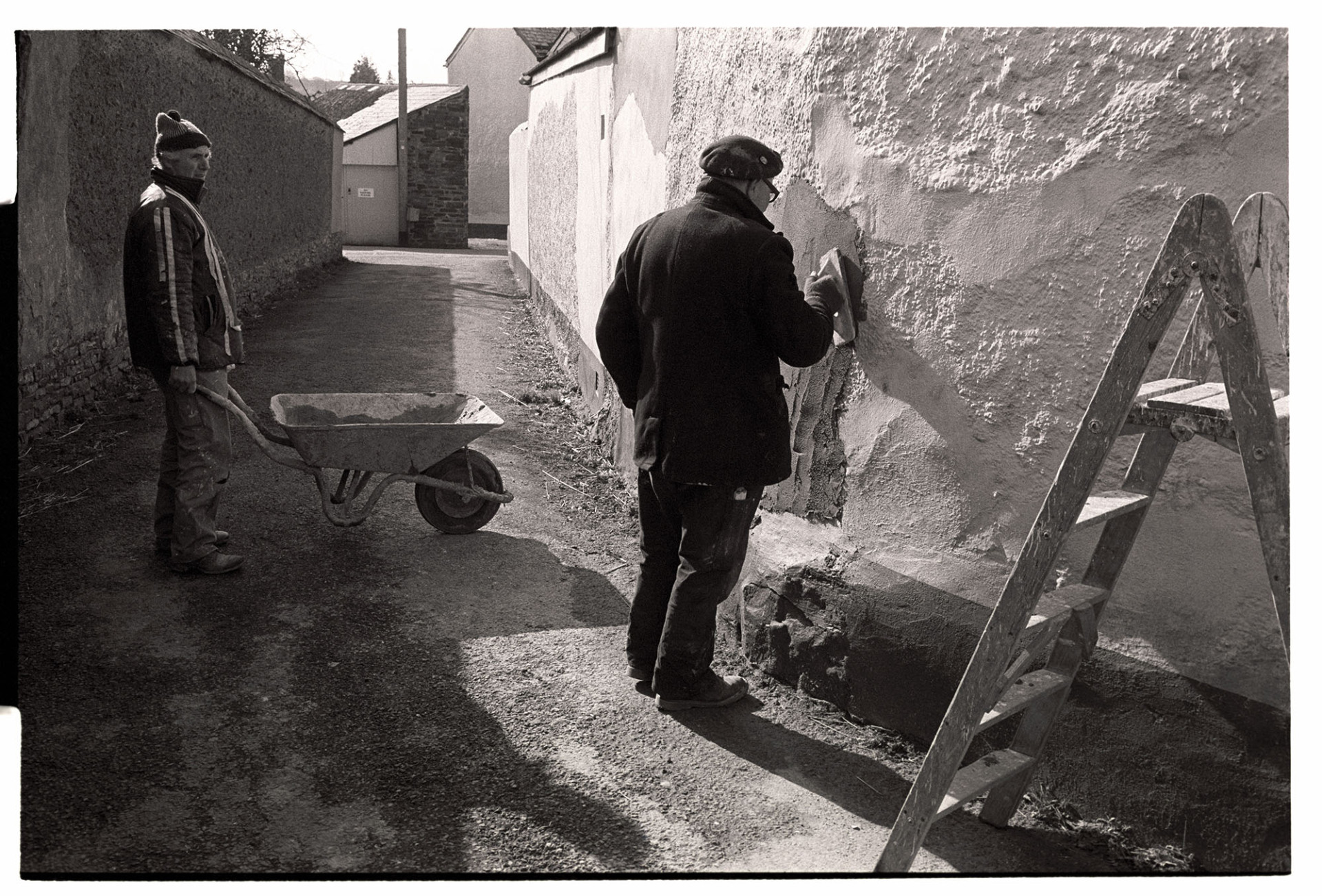 Men repairing cob wall. 
[Derek Marden and Clifford Palmer repairing the cob wall of a building in North Street, Dolton. One of the men is holding a wheelbarrow. A ladder is visible in the foreground.]
