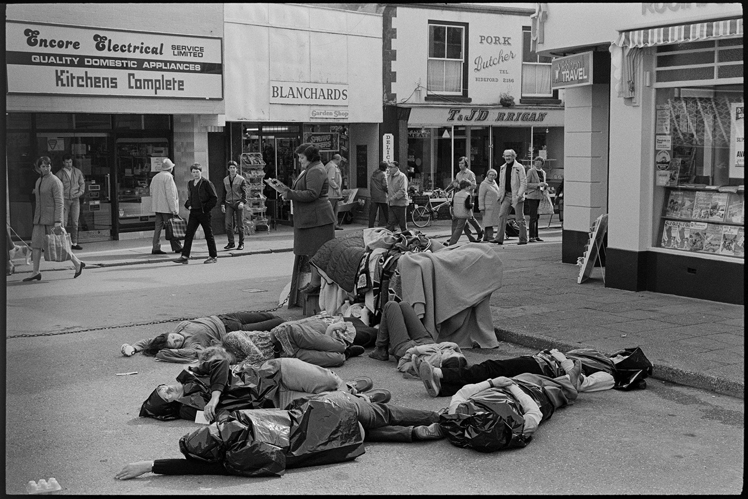 Women's peace movement demonstration, drama and singing songs, photographer. 
[A women's anti-nuclear demonstration in Mill Street, Bideford. The women are lying on the ground, whilst another woman reads from a booklet Shoppers walking past are looking at the demonstration. Shop fronts including Blanchards and Encore Electrical are visible on the opposite side of the street.]