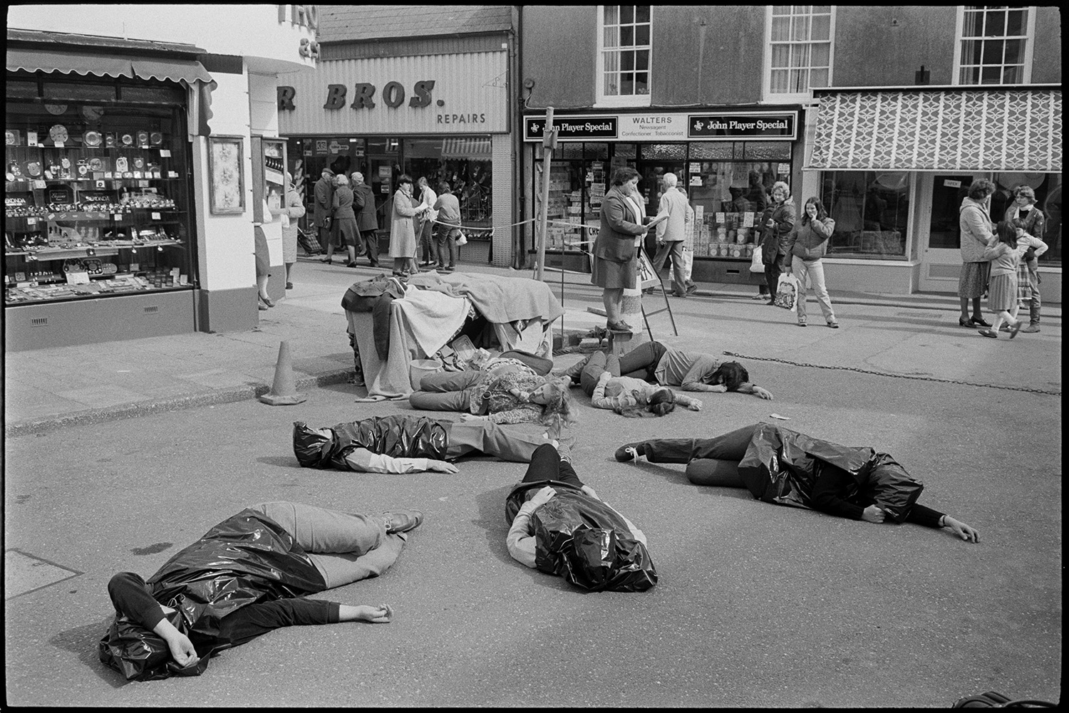 Women's peace movement demonstration, drama and singing songs, photographer. 
[A women's anti-nuclear demonstration in Mill Street, Bideford. The women are lying on the ground, whilst another woman reads from a booklet. Shoppers walking past are looking at the demonstration. The shop front of Walter's Newsagent is visible on the opposite side of the street.]