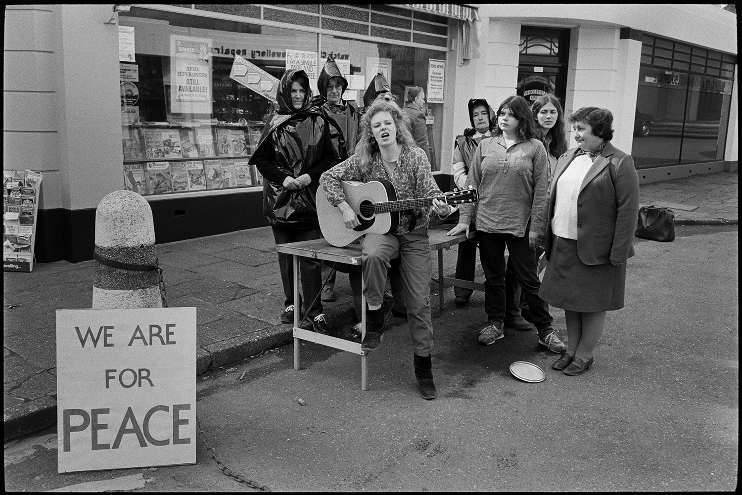 Women's peace movement demonstration, drama and singing songs, photographer. 
[A women's anti-nuclear demonstration in Mill Street, Bideford. One woman is playing a guitar with a small group gathered around her. A shop front can be seen in the background with a display of magazines in the shop window.]