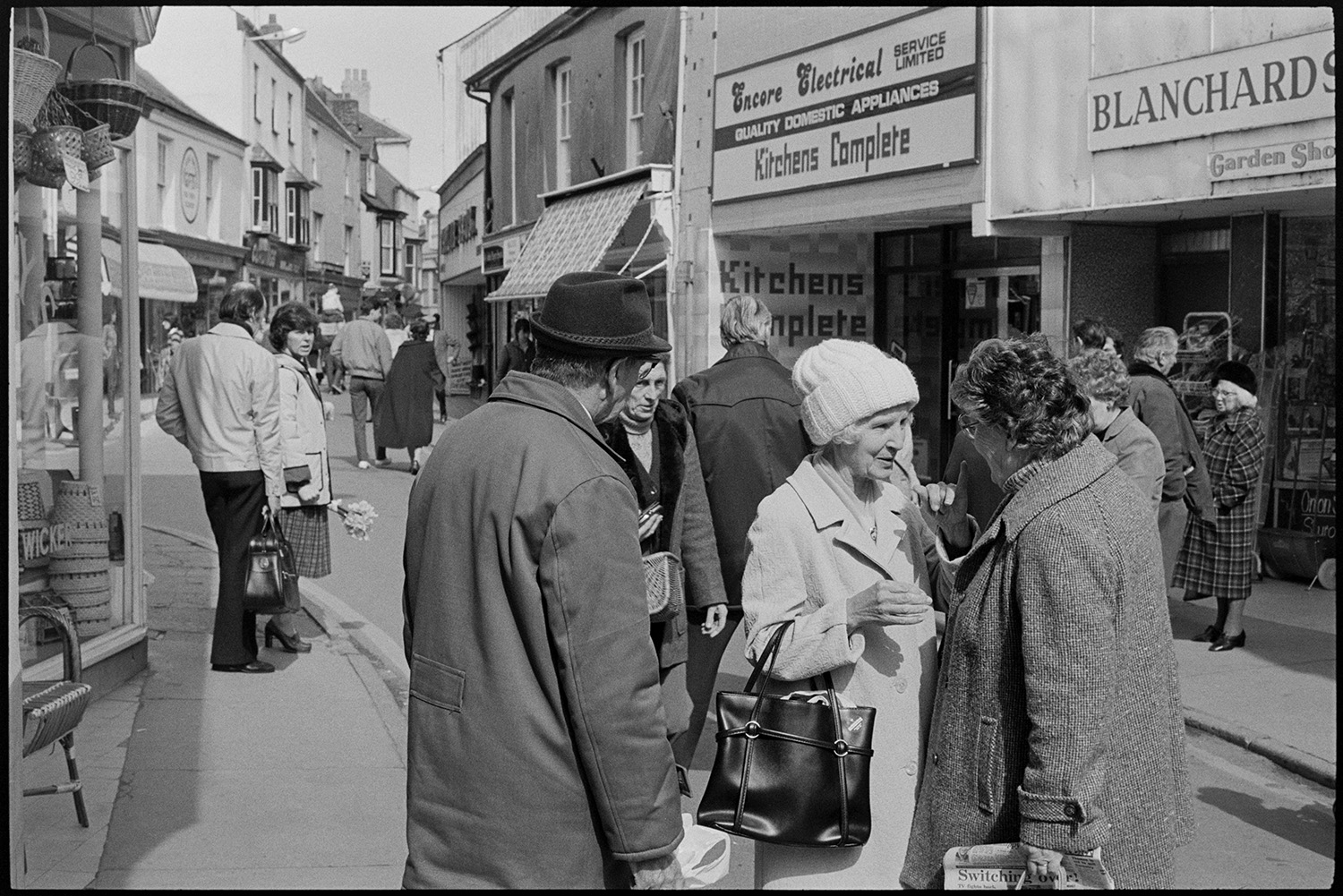People chatting in street, woman shopping. 
[Men and women chatting in Mill Street, Bideford outside 'Blanchards' and 'Encore Electrical' shop fronts. One of the women is wearing a woolly hat. Other shoppers can be seen in the street in the background.