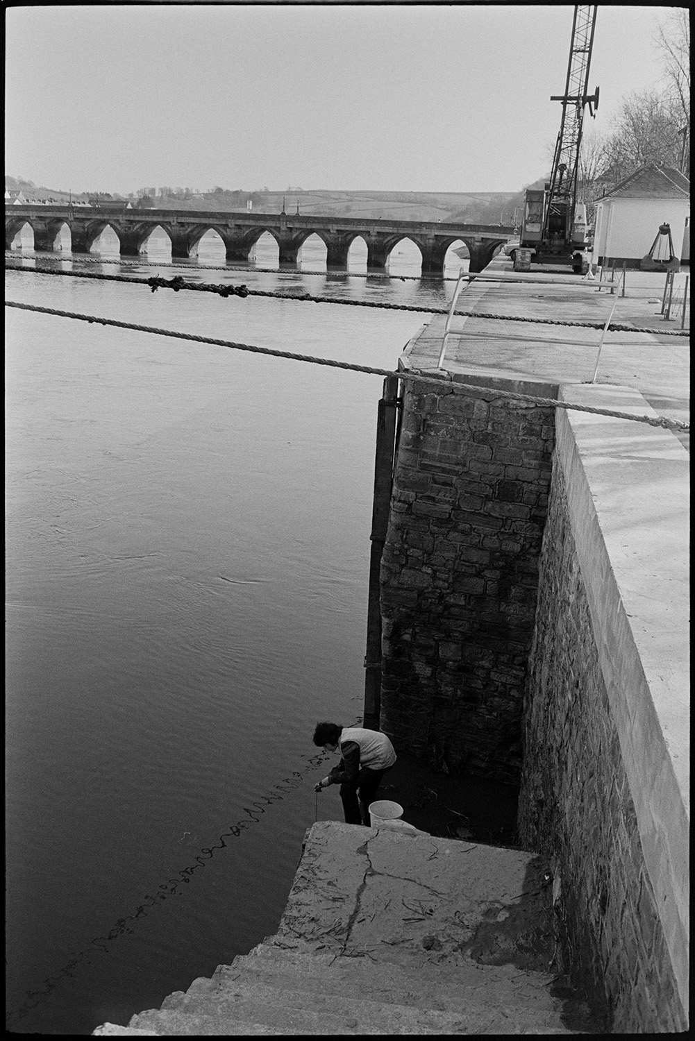 Boy fishing from quay near bridge. <br />
[A boy fishing at the bottom of steps leading down to the River Torridge at Bideford Quay. Bideford Long Bridge, also known as Bideford Old Bridge, can be seen in the background.]