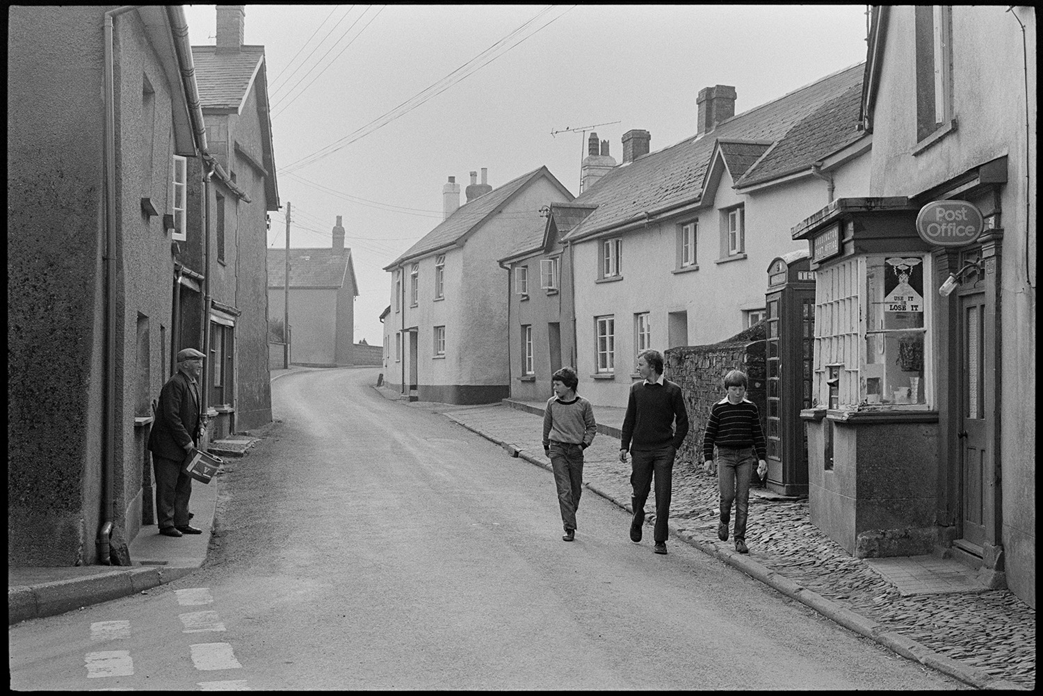 Street scene, early morning passers by, men chatting, front of Post Office, cobbles. <br />
[A man and two boys talking to another man on the opposite side of the street, as they walk past Burrington Post Office and telephone box in the early morning. The pavement is cobbled and houses can be seen further along the street.]