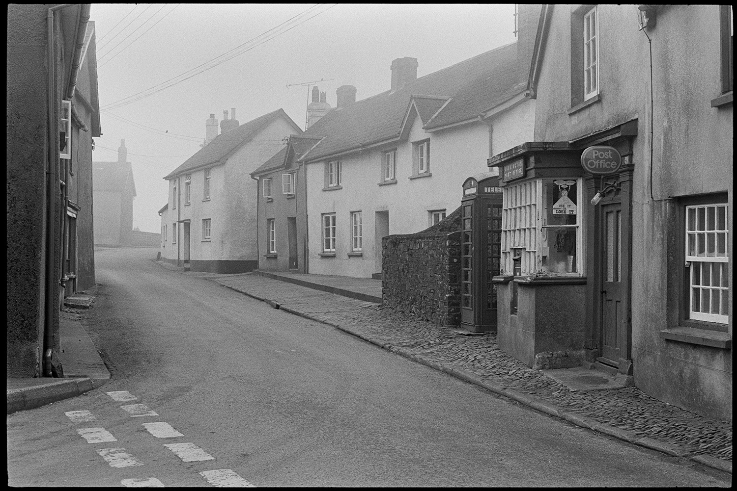 Street scene, early morning passers by, men chatting, front of Post Office, cobbles. 
[The street outside Burrington Post Office and telephone box in the early morning. The pavement outside the Post Office is cobbled.]