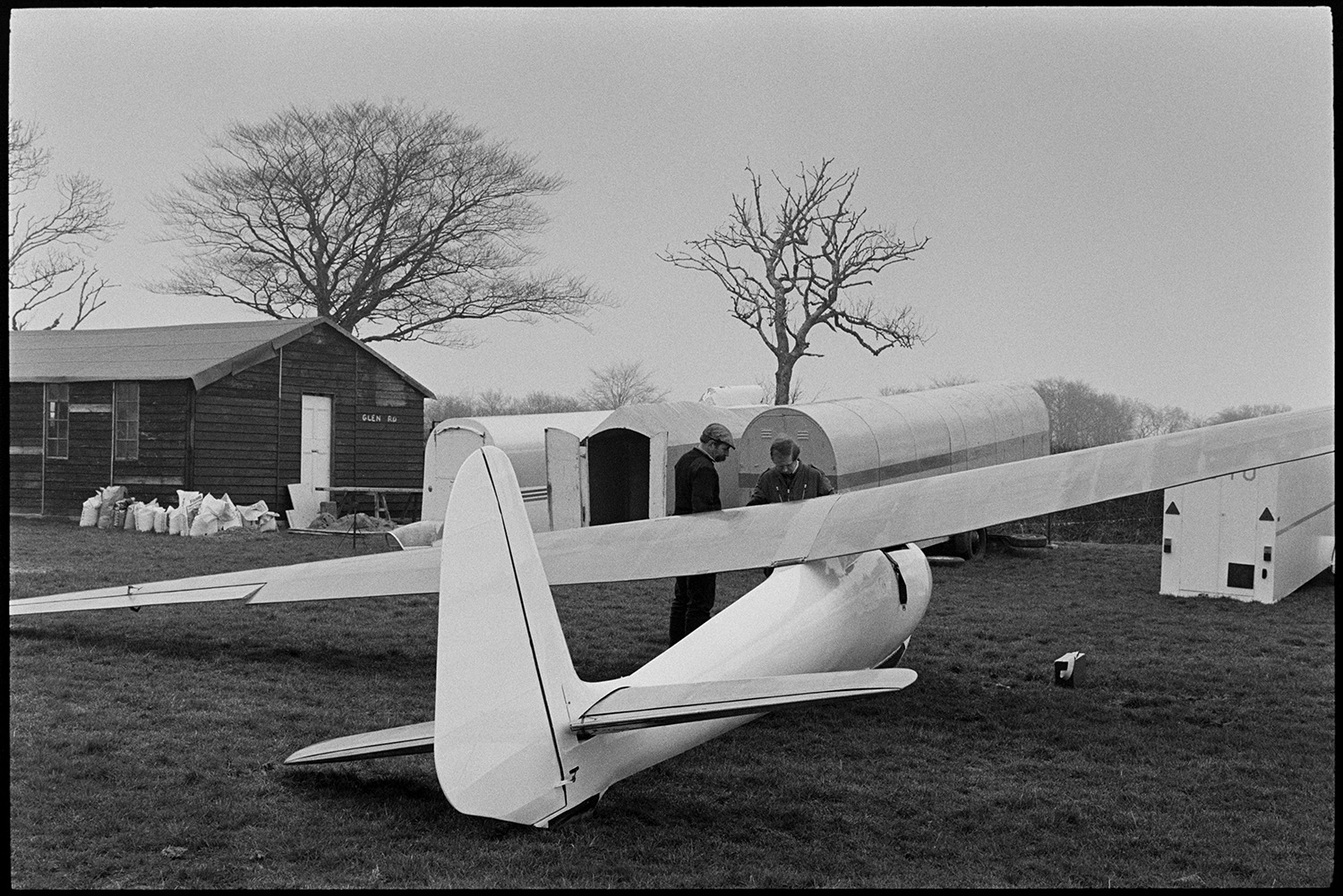 Glider at flying club. 
[Two men looking at a glider in a field at a flying club at Eaglescott, Burrington. Various trailers on wheels can be seen next to a wooden shed in the background.]