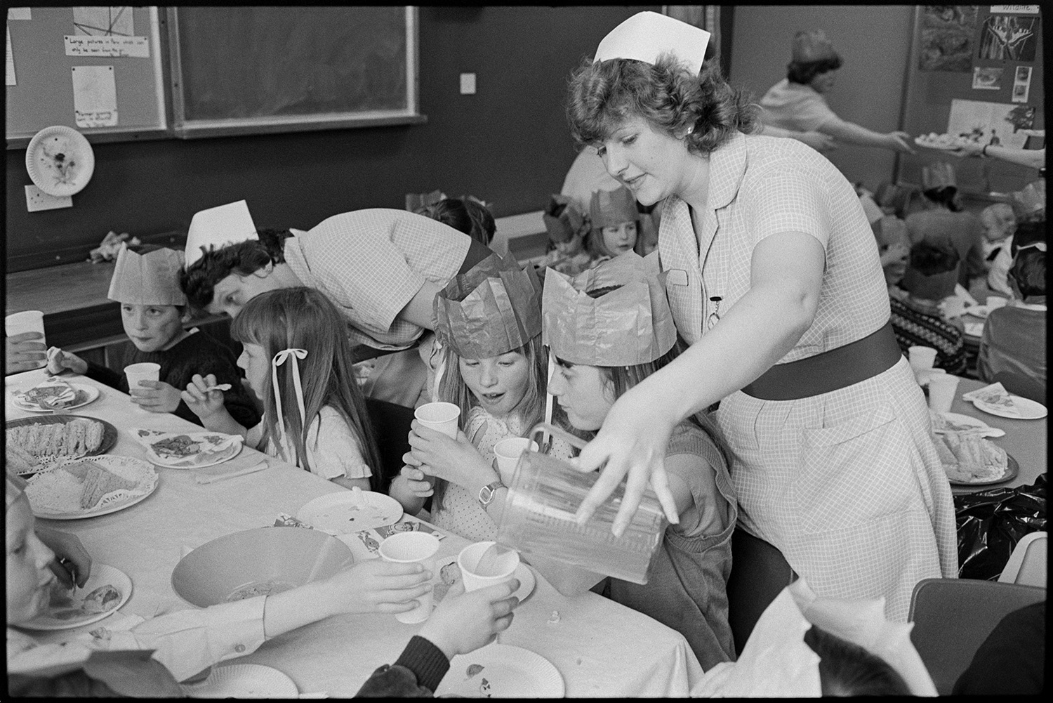 Party in children's ward of Hospital, food and drink served by nurses, buns, cakes. <br />
[Two nurses serving drinks and talking to children at a party on the Children's Ward at Barnstaple General Hospital. Some of the children are wearing paper hats. Cakes and sandwiches are laid out on the table.]