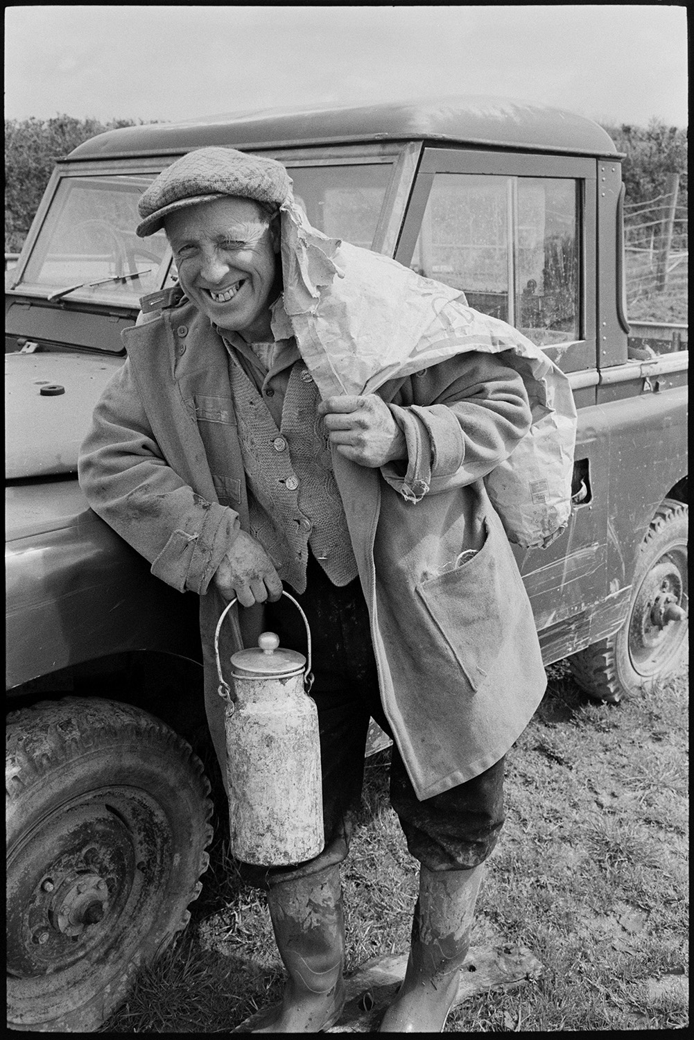 Farmer leaning against Land Rover, wearing cap, carrying bag of feed and milk can. 
[George Ayre leaning against a Land Rover in a field at Ashwell, Dolton. He is carrying a sack over his shoulder and a milk can or billy can in his other hand. He is also wearing a flat cap.]