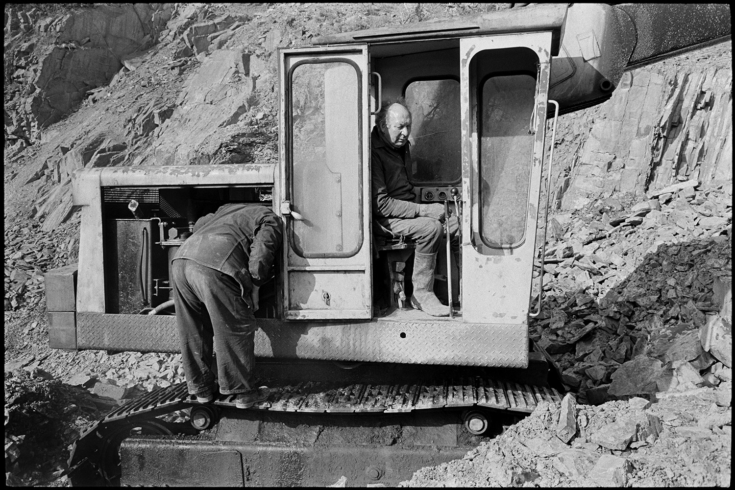 Men using mechanical shovel in quarry, excavator.
[Dennis Harris looking at the engine of an excavator at Newbridge Quarry, Dolton. He is stood on the caterpillar tracks of the digger. Harold Glover is sat in the cab of the excavator. Quarried stone can be seen in the background.]