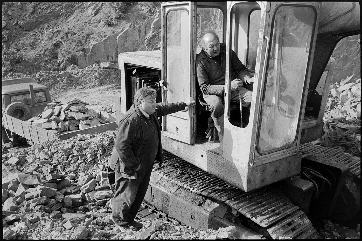 Men using mechanical shovel in quarry, excavator.
[Dennis Harris and Harold Glover working with an excavator at Newbridge Quarry, Dolton. Dennis Harris is leaning against the digger while Harold Glover is sat in the cab working the machine. A lorry loaded with stone is visible in the background.]