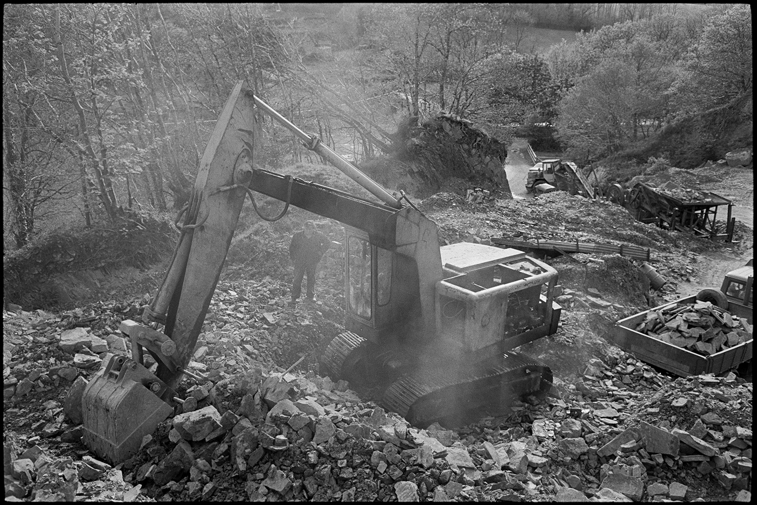 Men using mechanical shovel in quarry. Excavator.
[Dennis Harris and Harold Glover working with an excavator at Newbridge Quarry, Dolton. Dennis Harris is watching while Harold Glover works the digger to move a  pile of stone. A lorry is loaded with stone in the background and the entrance to the quarry can be seen amongst trees.]