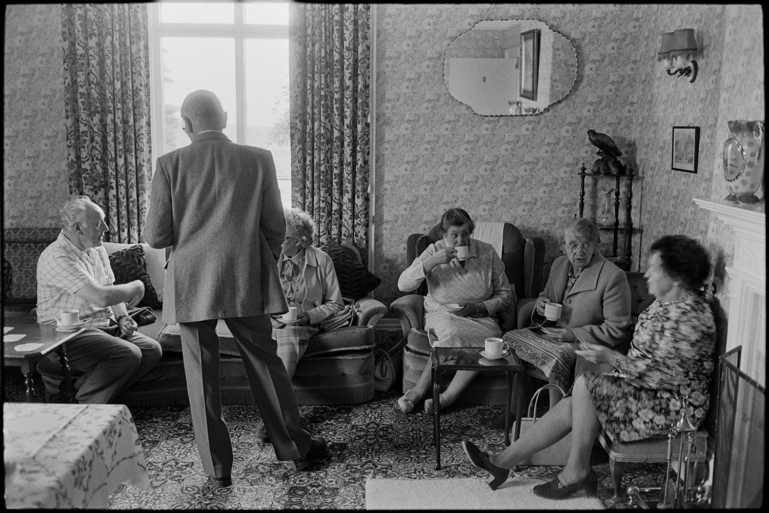 Women, people drinking tea in sitting room at fete.<br />
[Men and women sitting on armchairs and a sofa, drinking tea and chatting at a fete at the Old Rectory, Merton. The walls are covered with patterned wallpaper and a mirror is hung on the wall in the background.]