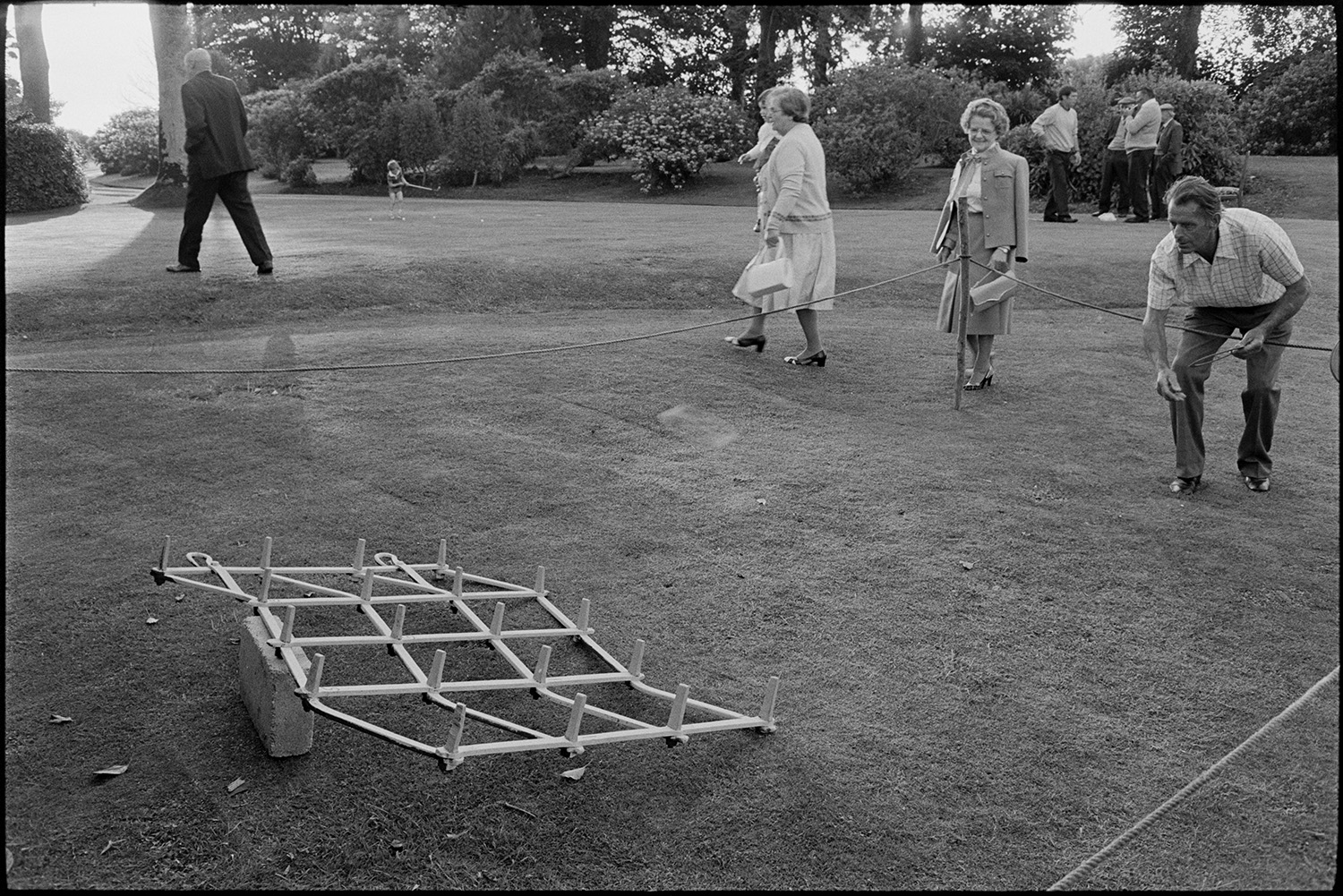 Sideshows at garden fete, bowls,  hoop la, billiards, golf. 
[A man playing hoop-la at a fete at the Old Rectory, Merton. A woman is watching him and in the background a group of men are talking by a trees and a child is playing with a golf club.]