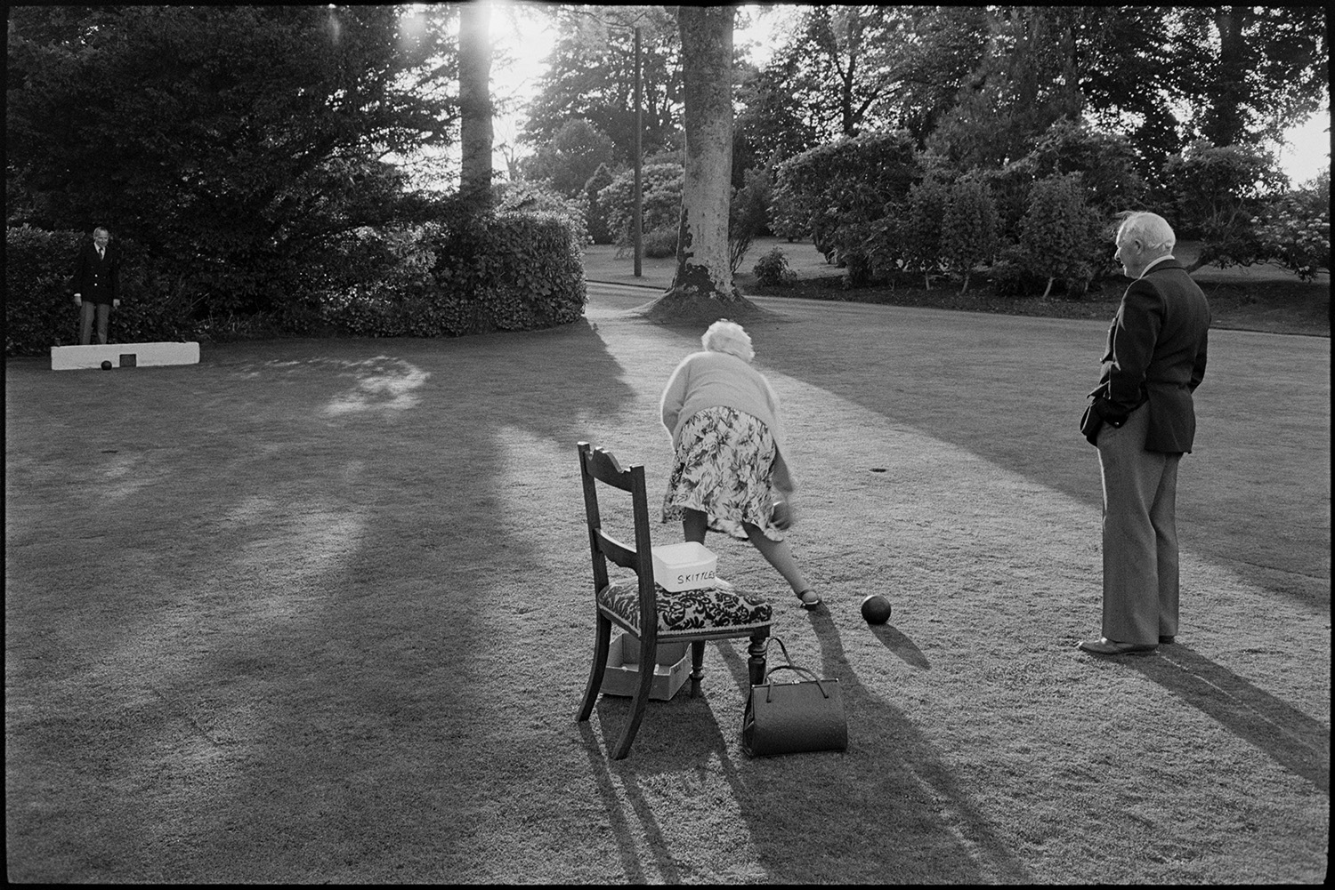 Sideshows at garden fete, bowls, hoop la, billiards, golf. 
[A man and woman playing bowls on the lawn at a fete at the Old Rectory, Merton. Shadows from trees are falling onto the grass.]
