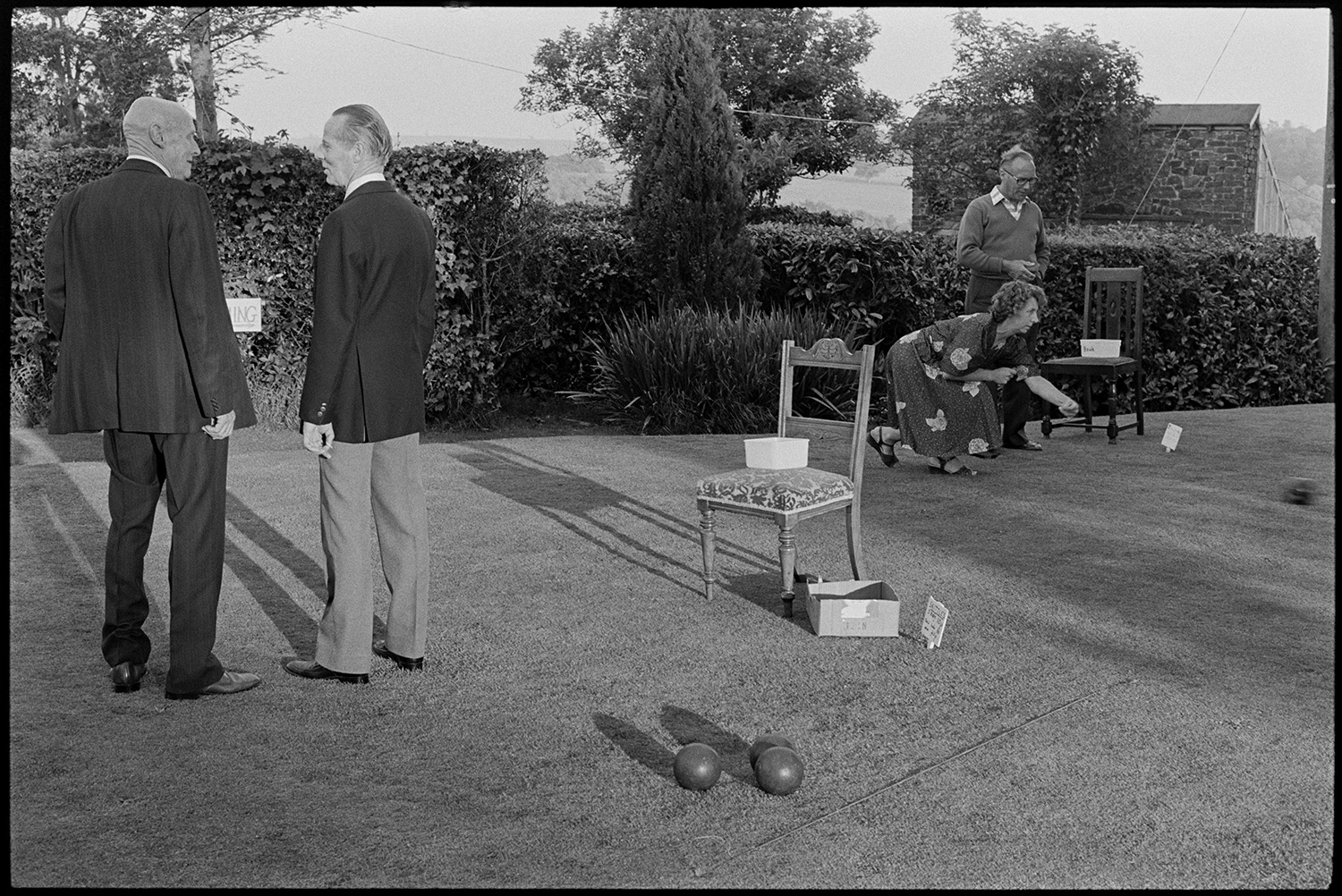 Sideshows at garden fete, bowls, hoop la, billiards, golf. 
[A man and woman playing bowls on the lawn at a fete at the Old Rectory, Merton. Two other men are talking in the foreground by a chair and another set of bowls. Shadows  from the chair and people are falling on the grass.]