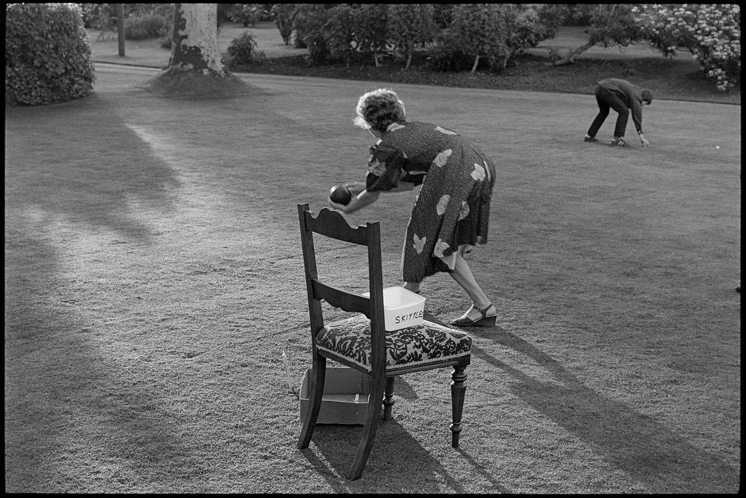 Sideshows at garden fete, bowls, hoop la, billiards, golf. 
[A woman playing bowls on the lawn at a fete at the Old Rectory, Merton. Shadows from trees are falling on the grass and a person is picking up golf balls in the background.]