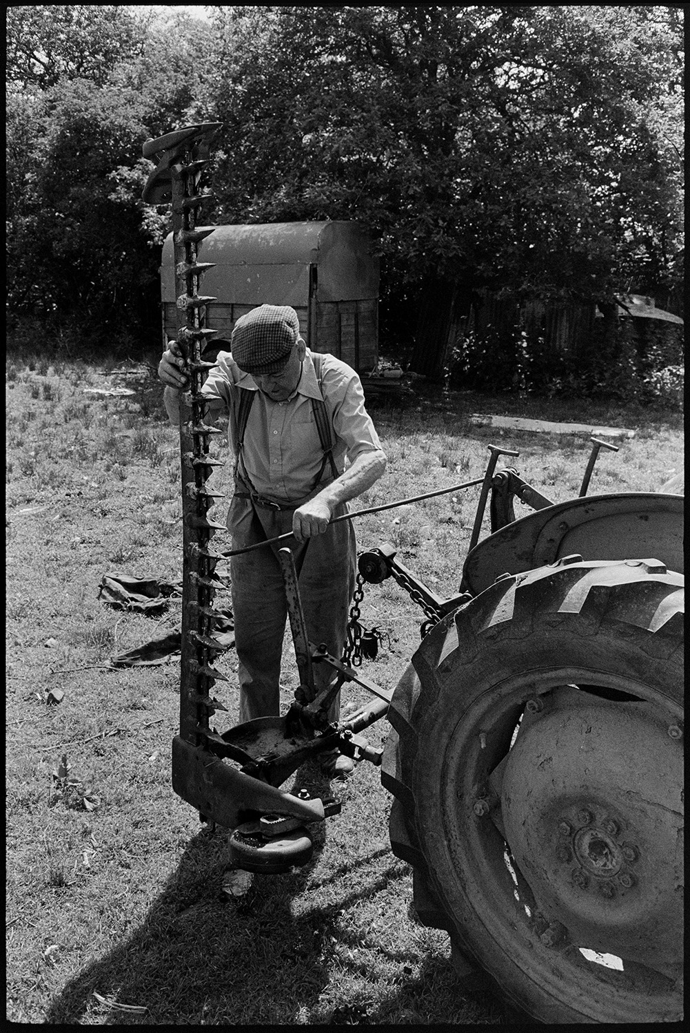 Farmer shearing sheep under tree. Woman farmer bundling up fleeces.
[Cyril Bennett folding up a grass cutting machine attached to a tractor in a field at Cuppers Piece, Beaford. A trailer under a hedge is visible in the background.]