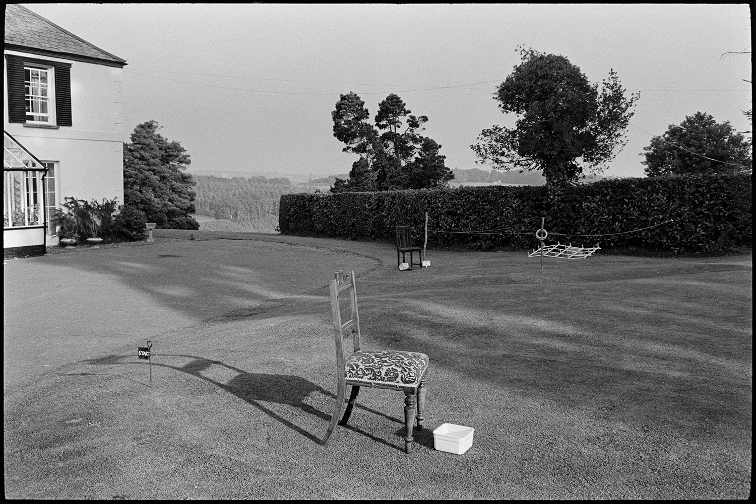 Jumble stalls at garden fete people buying.
[A chair set out for a game and a game of hoop la on the lawn at a fete at the Old Rectory, Merton. Shadows from trees and the chair are falling across the lawn.]