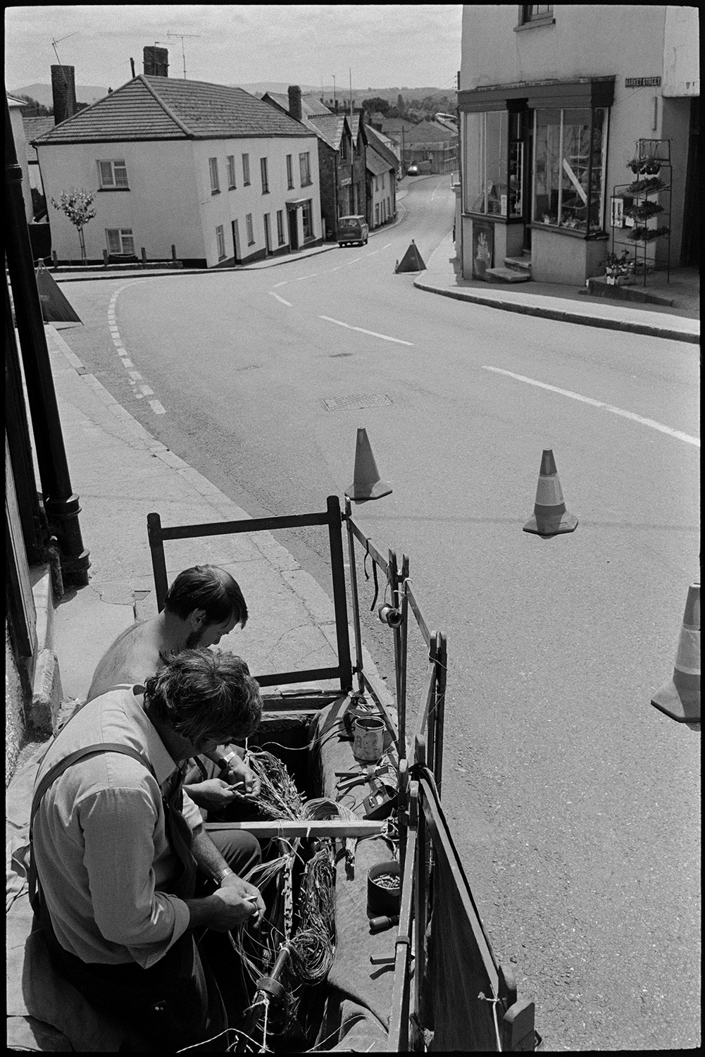 Electrical engineers working in pavement manhole in street.
[Engineers working on wires in open manholes in the pavement on Market Street in Hatherleigh. A shop front can be seen further down the street and traffic cones are in the road.]