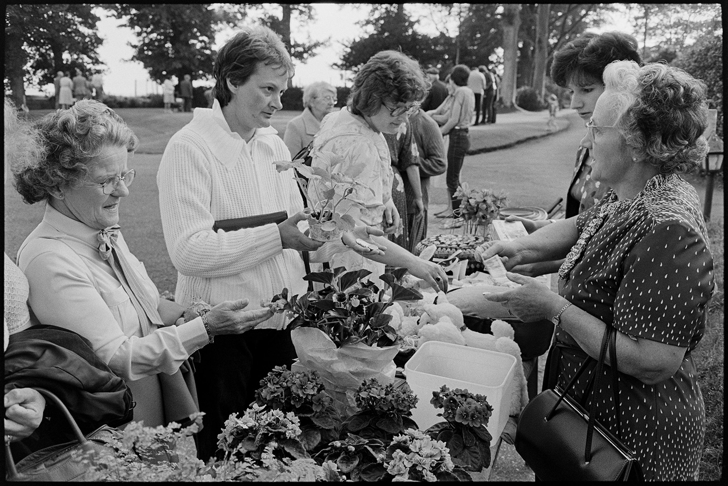 Jumble stalls at garden fete people buying.
[Men and women talking to a woman running a plant stall at a fete at the Old Rectory, Merton. Soft toys are also for sale on the stall.]
