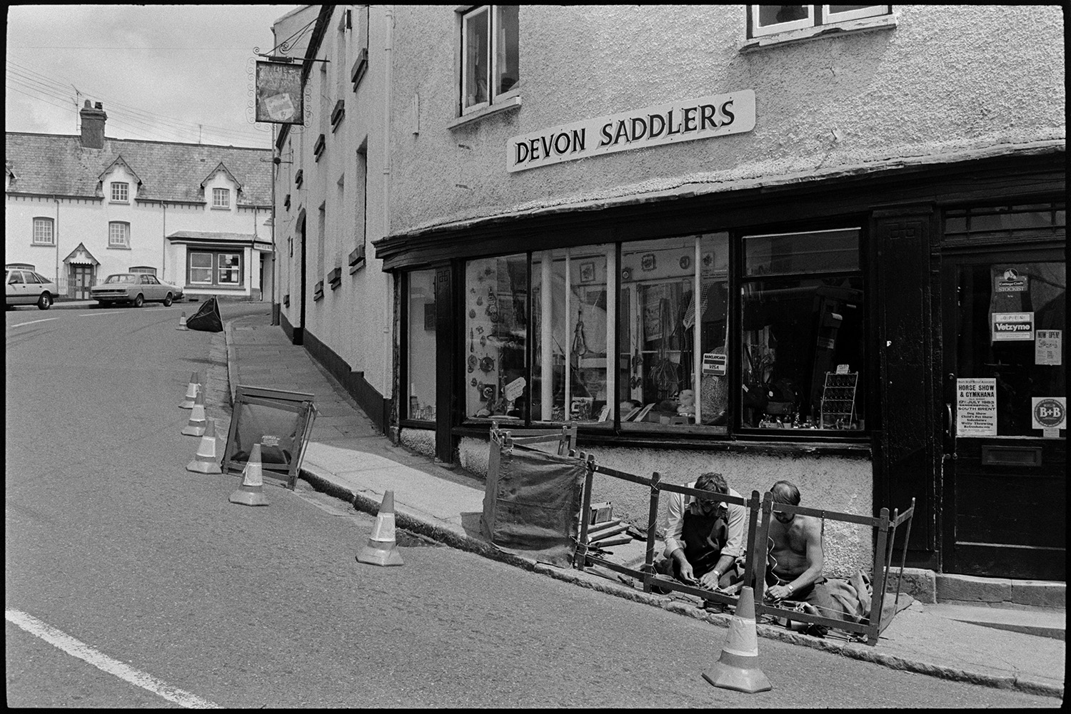 Electrical engineers working in pavement manhole in street.
[Engineers working on wires in open manholes in the pavement outside the Devon Saddlers shop in Market Street, Hatherleigh. Traffic cones are positioned by the pavement.]