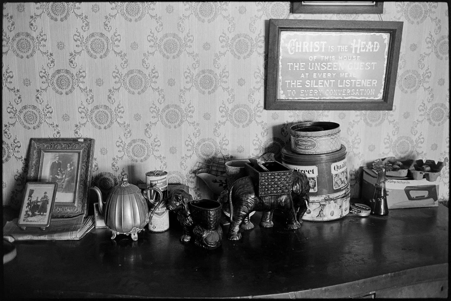 Farmhouse interior, side table, harmonium, electric fire.
[Ornaments, including a dog and elephant, framed photographs, tins, egg boxes and an electric iron box on a side table in the Ford family house at Venton, near Hollocombe. A religious framed motto is hung on the wall behind the table.]