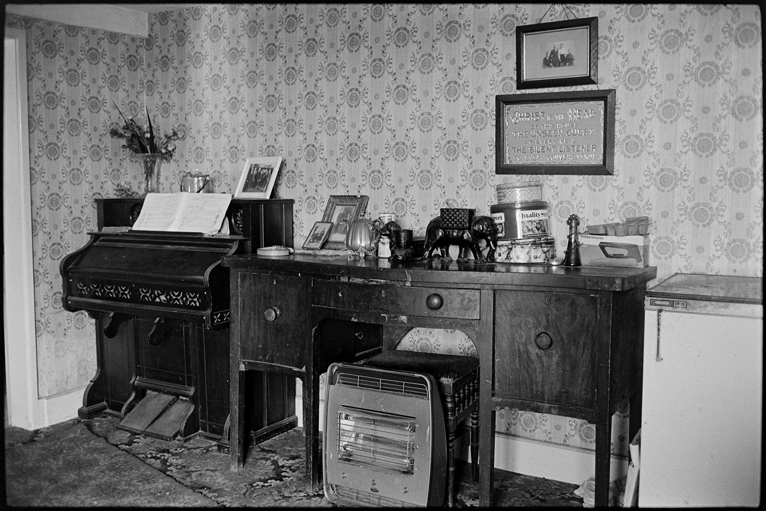 Farmhouse interior, side table, harmonium, electric fire.
[Ornaments, tins and framed photographs on a side table next to a Harmonium, a freezer and an electric fire in the Ford family house at Venton, near Dolton. A religious framed motto and family picture are hung on the wall in the background. The wall is covered with patterned wallpaper.]
