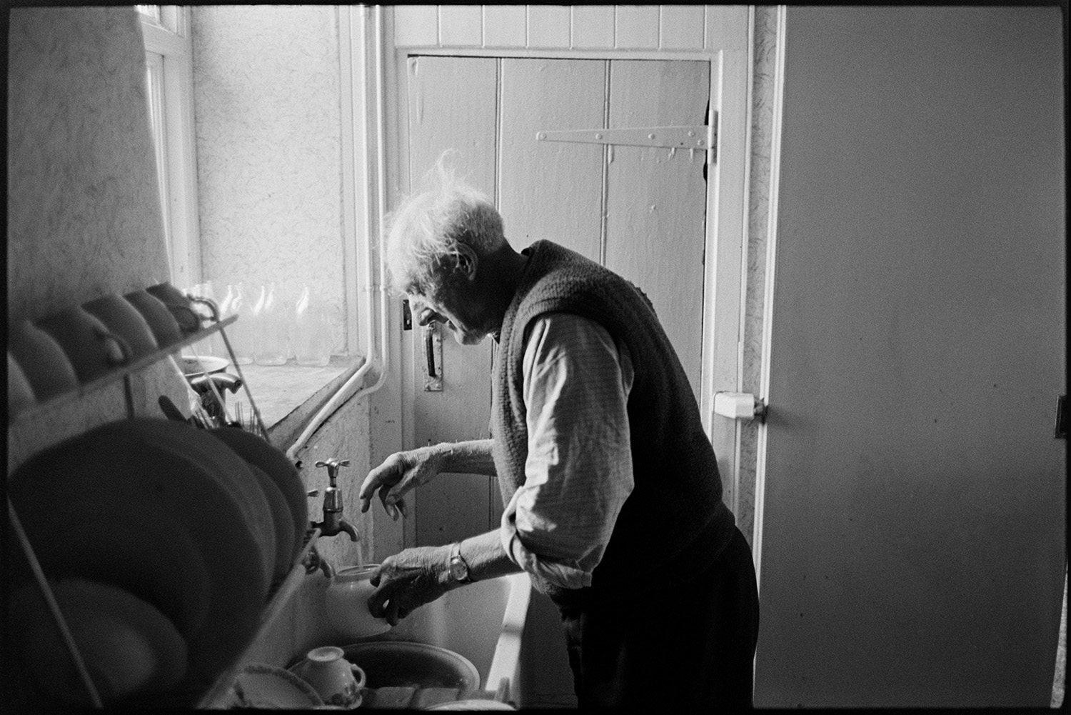 Farmer making pot of tea in kitchen, leaning out of back door, cat. 
[Arthur Ford filling a teapot with water from a tap in his kitchen at Venton, near Dolton. Plates and cups are stacked on a drying rack next to the sink.]