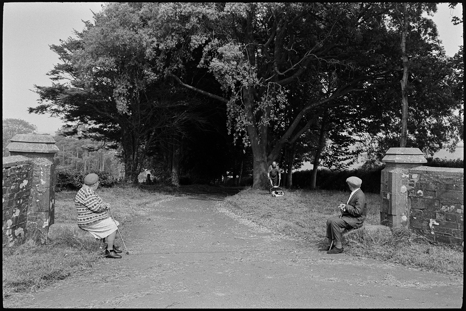 Elderly couple resting at gateway to country house. 
[Bessie Bright and Tom Bright sat on stones at the gateposts to Yatton Court, Beaford. They are watching a man mowing the grass at the entrance to the country house.]