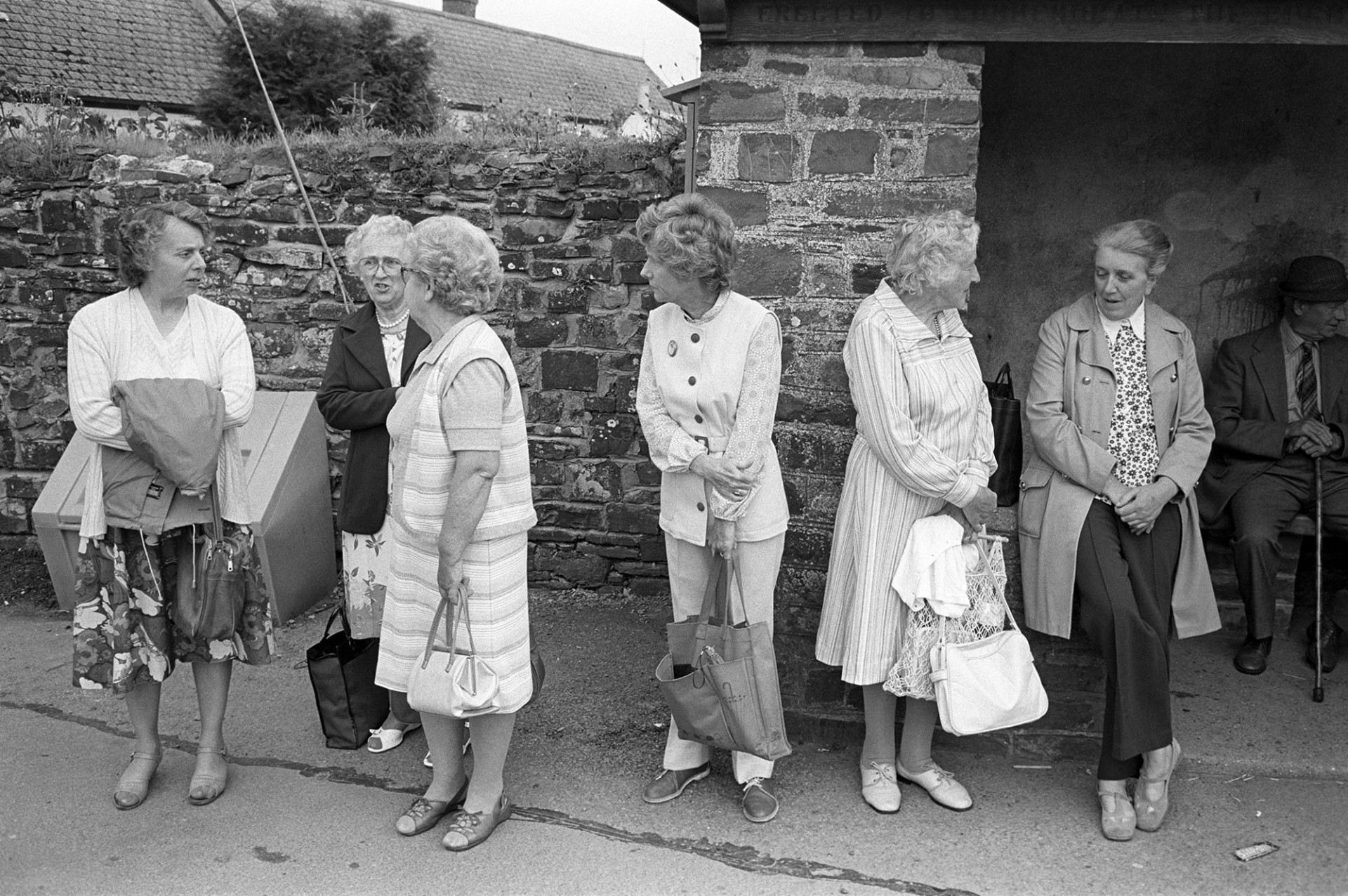 Women waiting for bus on Friday market day, dresses for summer. 
[Women including Mrs Down, Mrs Heard, Mrs Pickard and Mrs Ayre waiting at a bus shelter in Dolton for the Friday bus to go to a market. Some of the women are wearing summer dresses.]