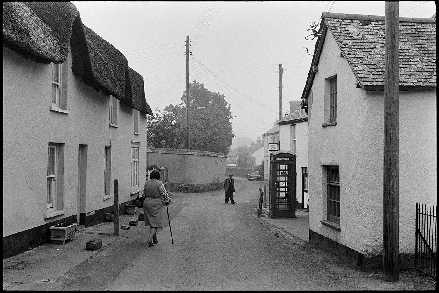 Village street, car, man posting letter, dog, telephone kiosk. 
[A man and a woman using a walking stick walking past thatched cottages in Fore Street, Dolton. A telephone box in on the other side of the street.]