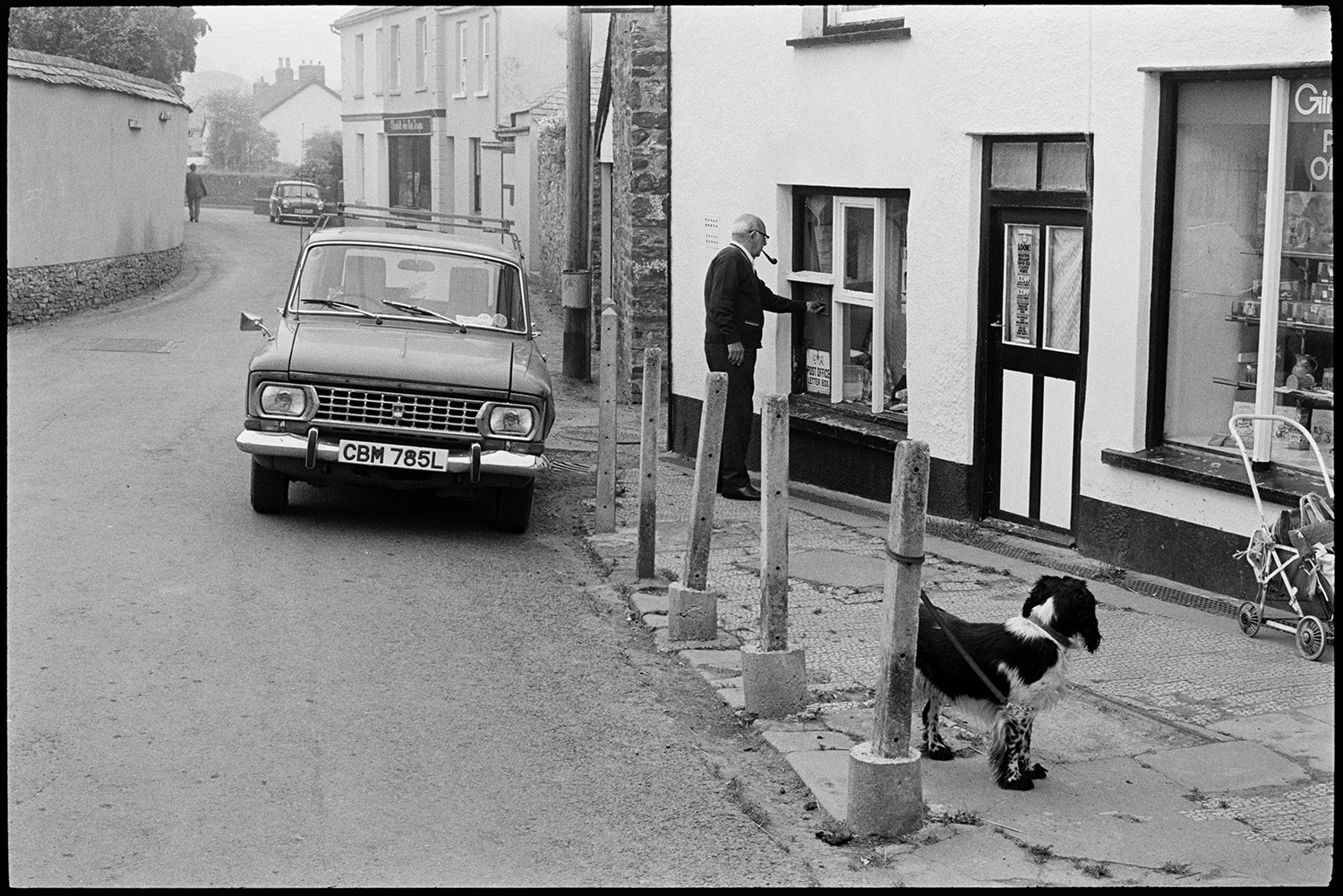 Village street, car, man posting letter, dog, telephone kiosk. 
[A man posting a letter in a post box in the window of Dolton Post Office. A pram, a dog tied to a post and a car are waiting outside the shop front.]