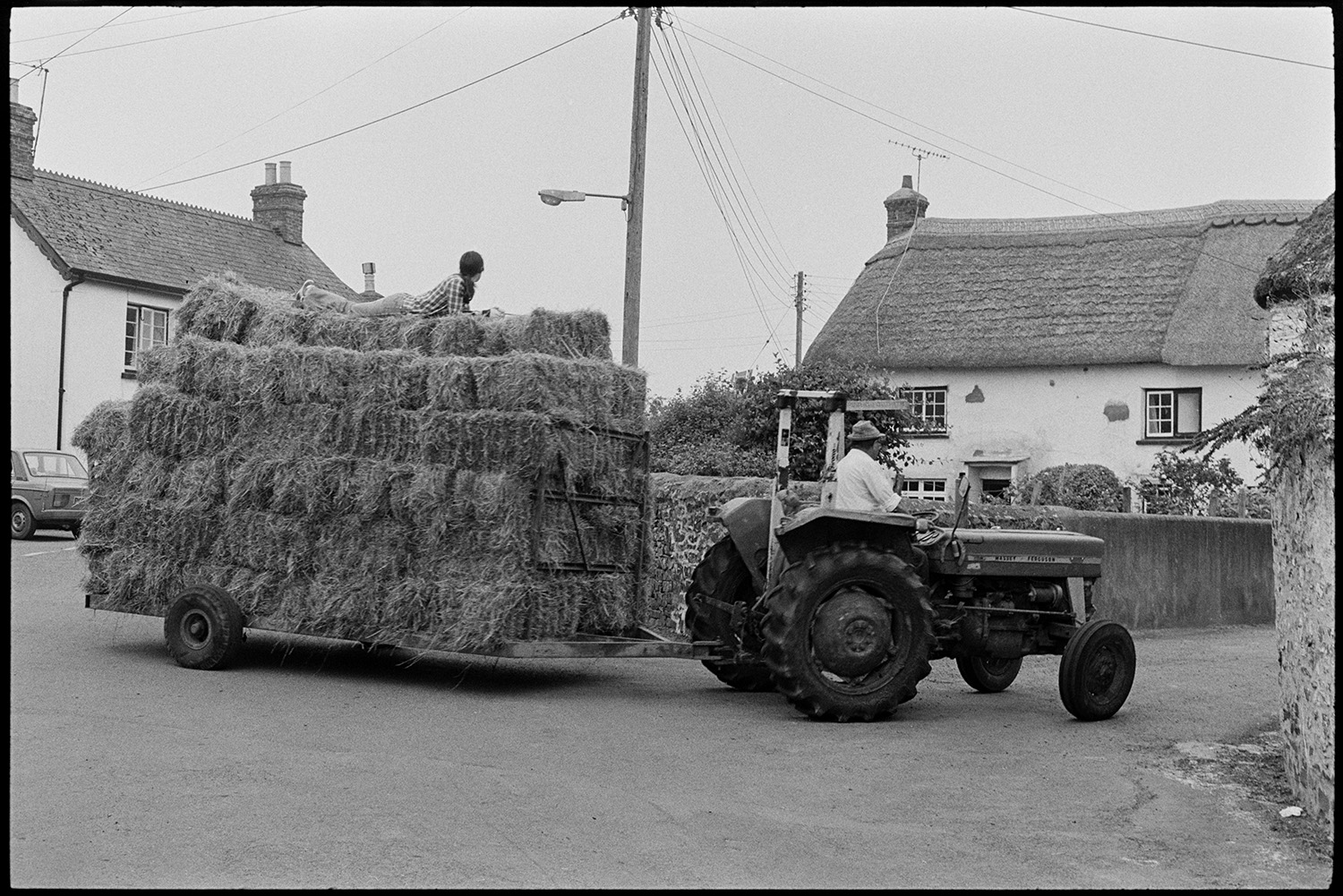 A man driving a tractor and trailer loaded with hay through a street, possibly in Dolton. A person is laying on top of the hay.