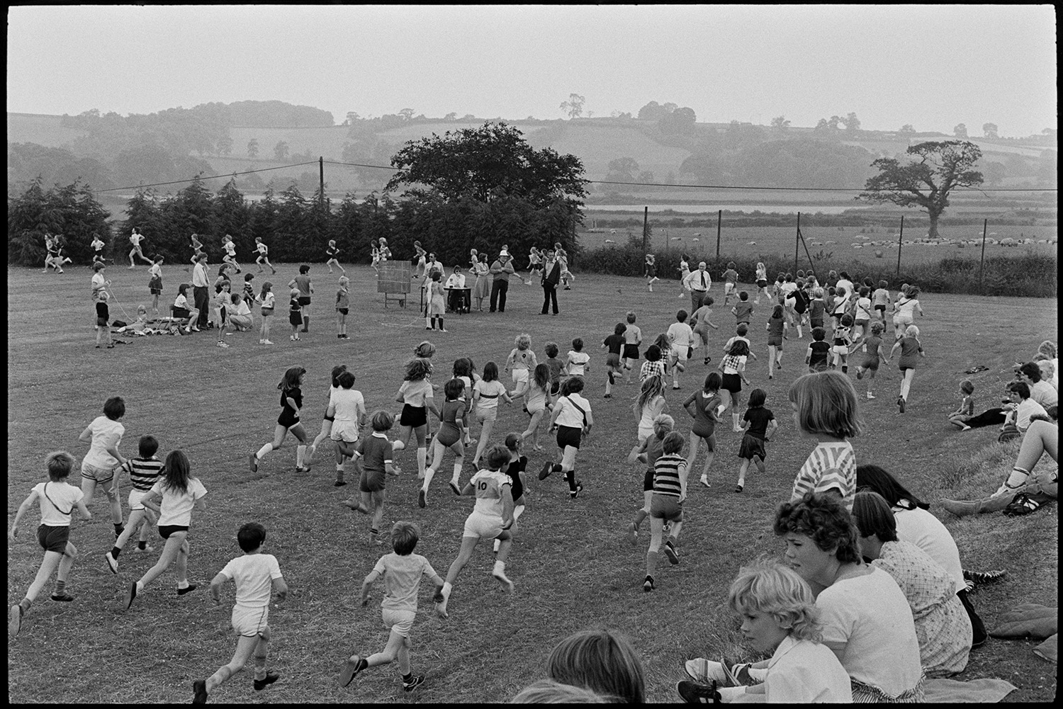 School sports day, races and prizes being given. 
[Children running in a race at Winkleigh School sports day on the school field. Parents and young children are watching the race from a grassy bank on the edge of the field.]