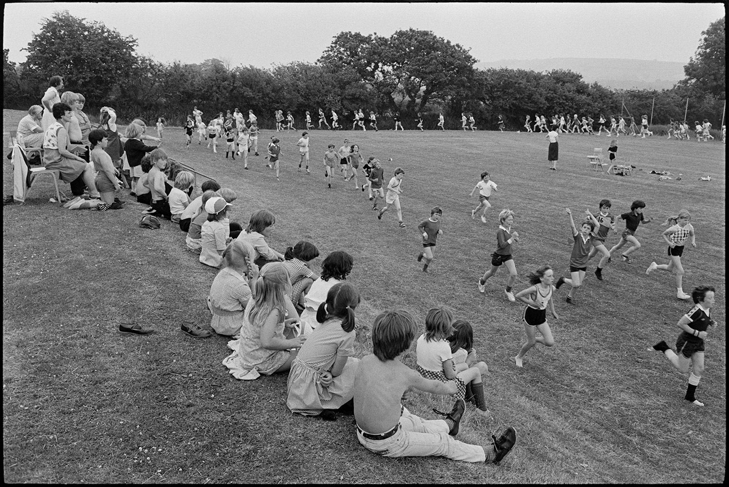 School sports day, races and prizes being given. 
[Parents and young children watching children running in a race at Winkleigh School sports day on the school field. They are sat on a grassy bank on the edge of the field to watch the race.]