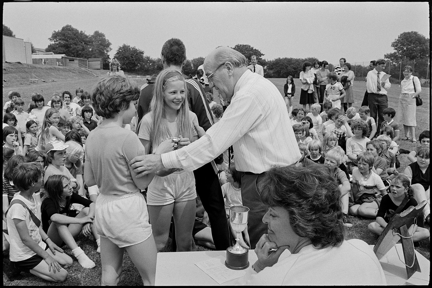 School sports day, races and prizes being given. 
[A man handing a trophy to two children at Winkleigh School Sports Day on the school field. Other schoolchildren and teachers are gathered around for the prize giving.]