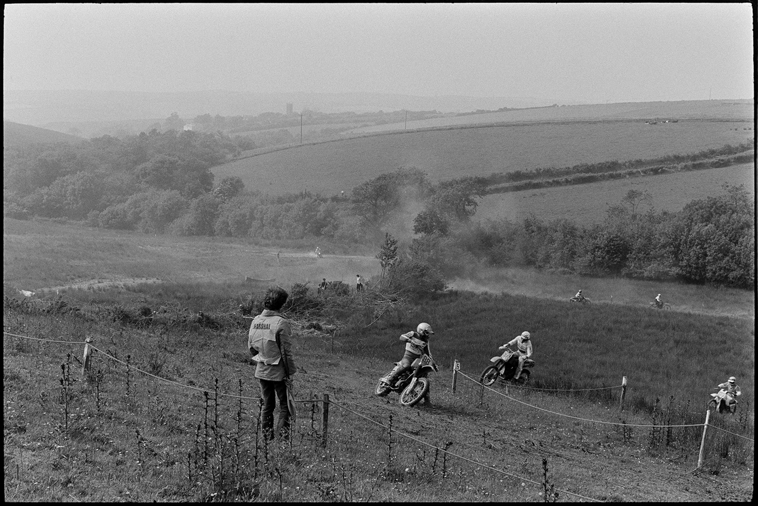 Motorcycle scramble. 
[A marshal watching motorcyclists race around a track at a motorcycle scramble in a field at Torrington.]