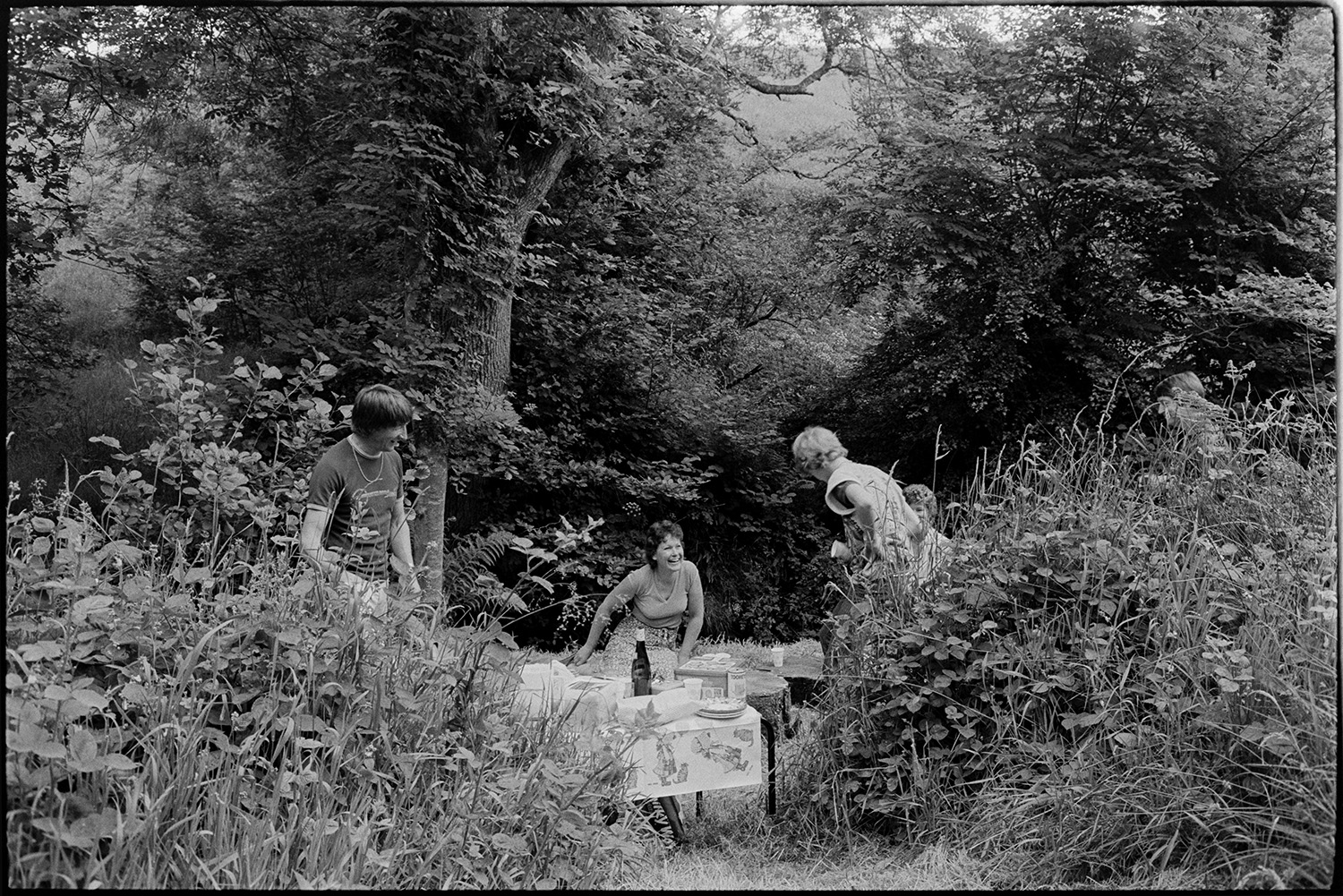 People picnicking in wood beside stream. 
[Women and a man having a picnic in a small clearing by a stream in Dolton Wood. Food and drink are laid out on a small table and tree stumps.]
